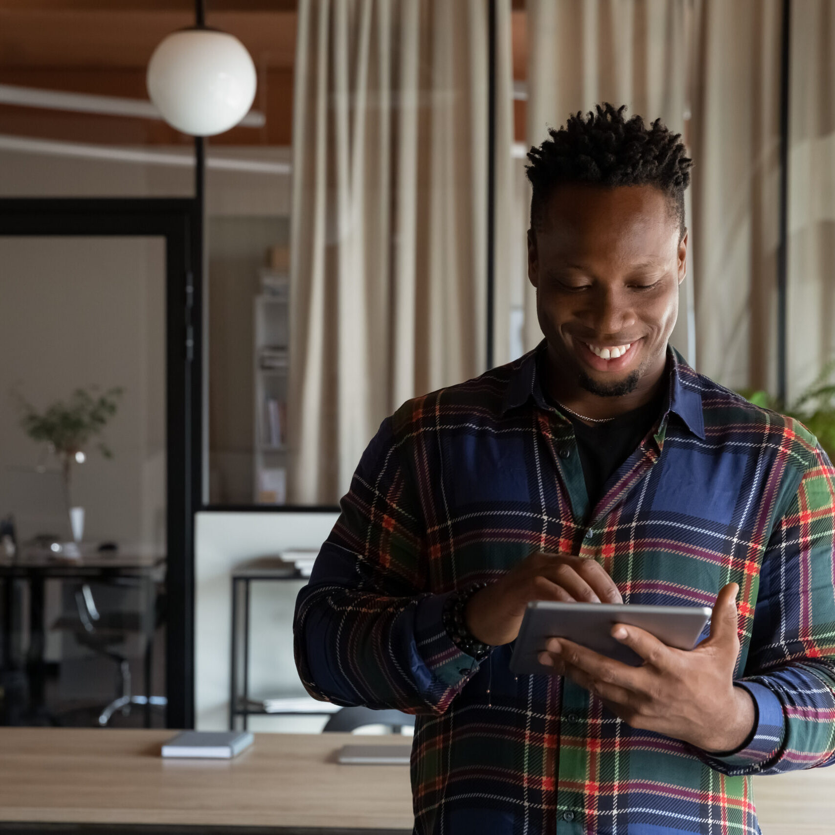 Happy millennial self employed professional using tablet in office. Young African American hipster guy, student, employee using virtual app, online service on pad computer, chatting, browsing