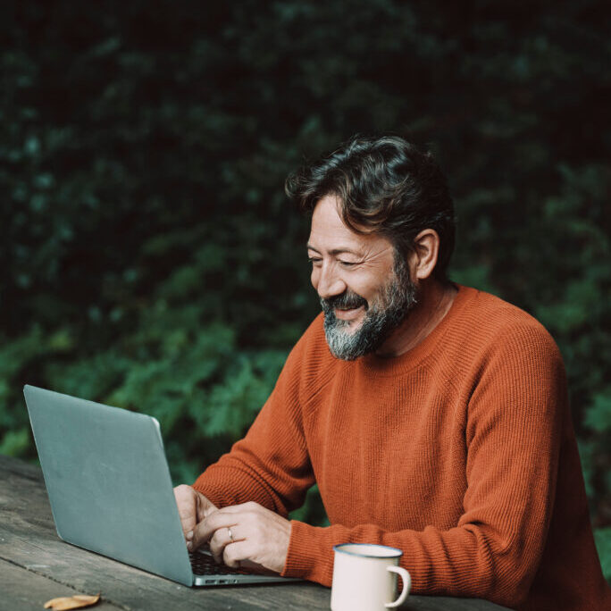 Happy free adult man work outdoors on a table with nature in background - concept of digital nomad modern online lifestyle people - mature male with beard use laptop computer in the woods smiling and enjoying